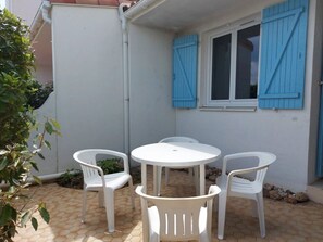 A patio with a round table and four white plastic chairs, a window with blue shutters, and a white wall.