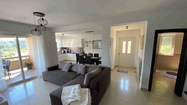 A spacious living area with a dark-colored sofa and a white throw blanket. A kitchen is visible in the background with white cabinets and a dining area. - Holiday apartment with balcony (Kampor)