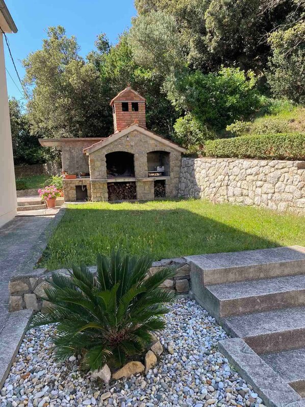 A stone-built outdoor kitchen with a chimney, surrounded by a stone wall and lush greenery.