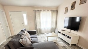 Un salon comprenant un canapé d’angle, une table basse, un téléviseur posé sur un meuble blanc et une grande fenêtre avec des rideaux.