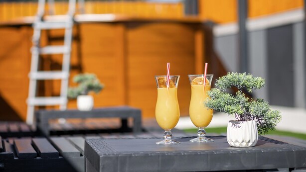 Deux verres d’une boisson à l’orange sont posés sur une table, avec une échelle et une petite plante en pot en arrière-plan.