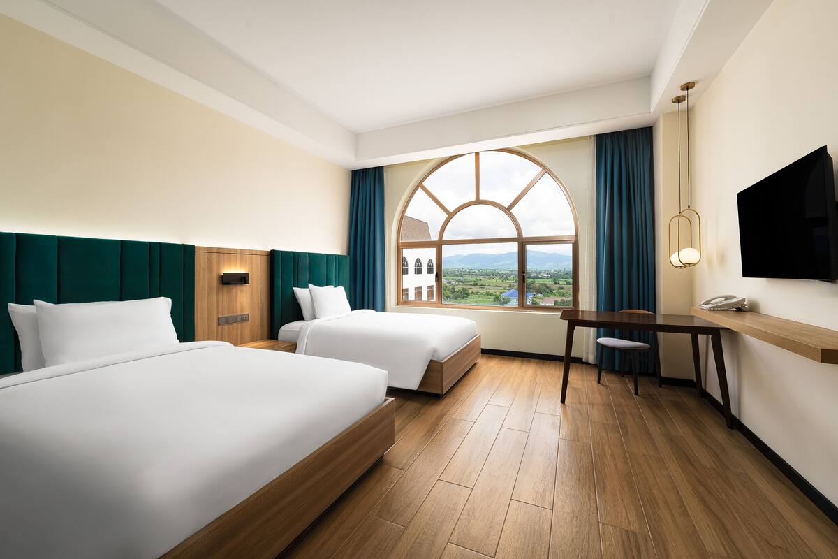 A hotel room with two beds, a wooden desk, a TV, and a large window with a view of the countryside.