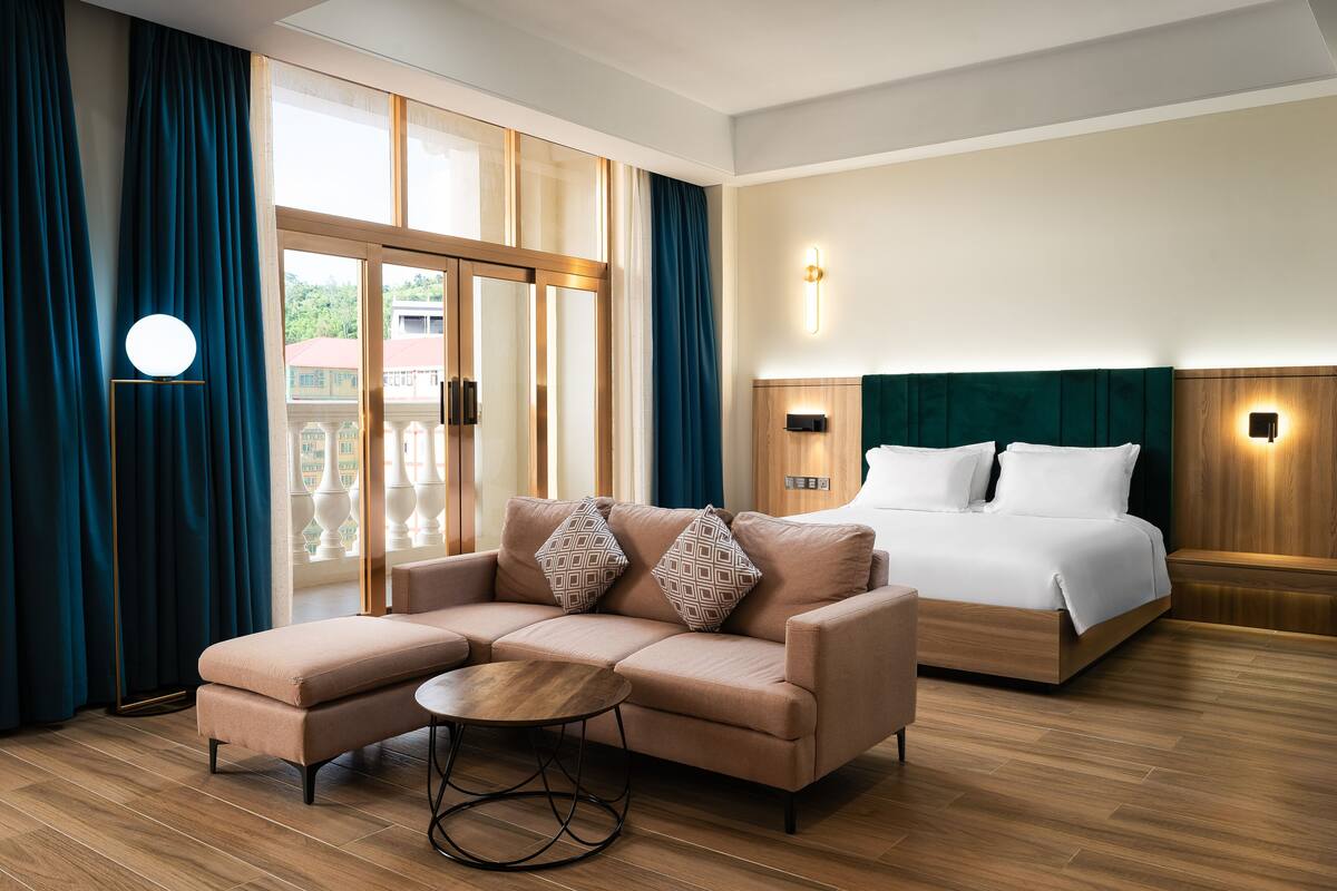 A modern hotel room with a sectional sofa, a round coffee table, a bed with a wooden headboard, and a view through a large glass door.