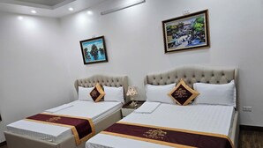 A hotel room with two beds, each with a headboard, white bedding, and a maroon runner. There are two framed pictures on the walls, a nightstand with a lamp, and a wooden floor. - Hoàng Gia Hotel Royal (Le Ho)