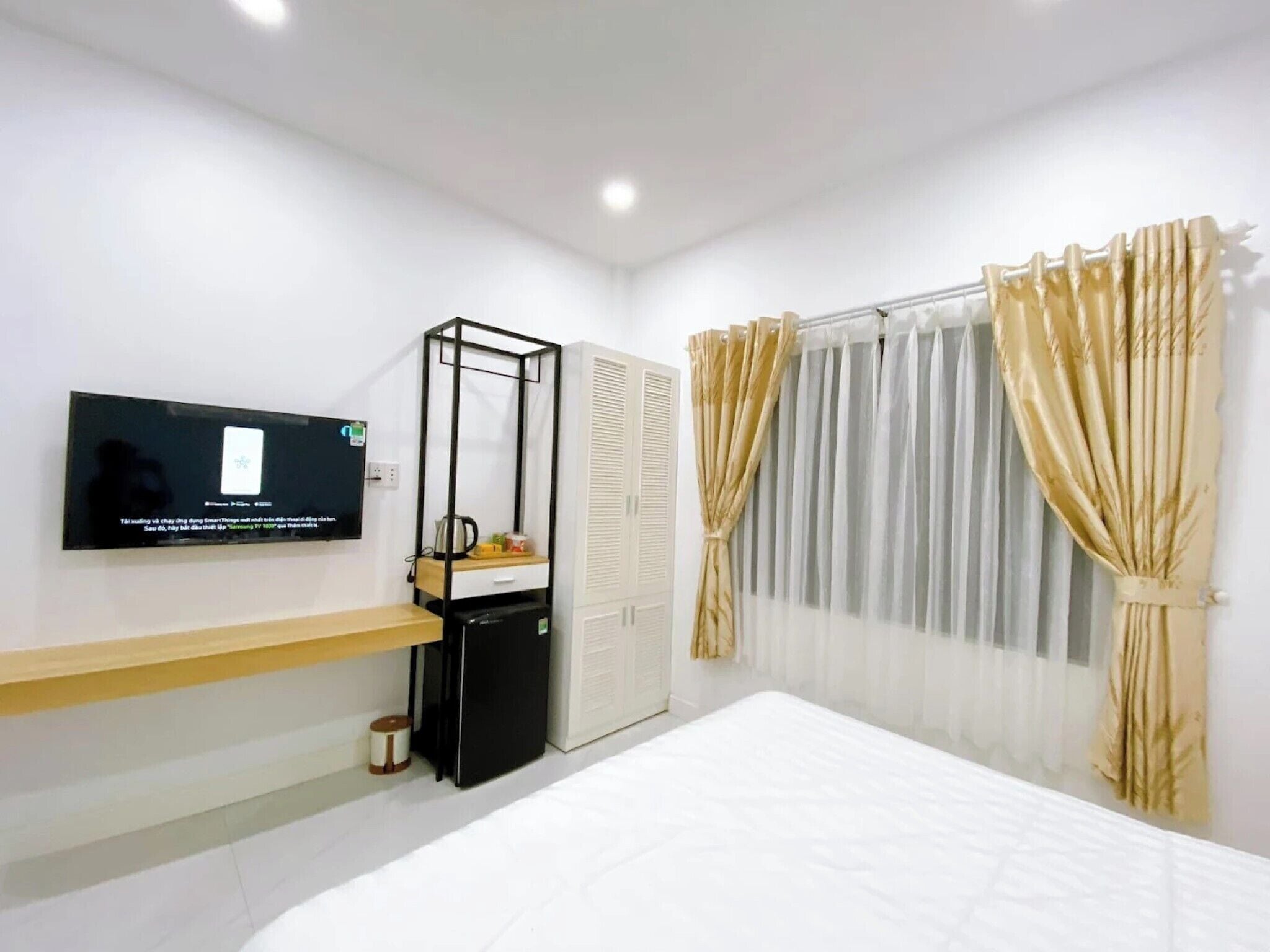 A modern hotel room with a bed, a TV mounted on the wall, a small desk with a chair, a wardrobe, and a window with curtains.