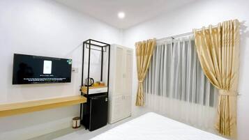 A modern hotel room with a bed, a TV mounted on the wall, a small desk with a chair, a wardrobe, and a window with curtains.