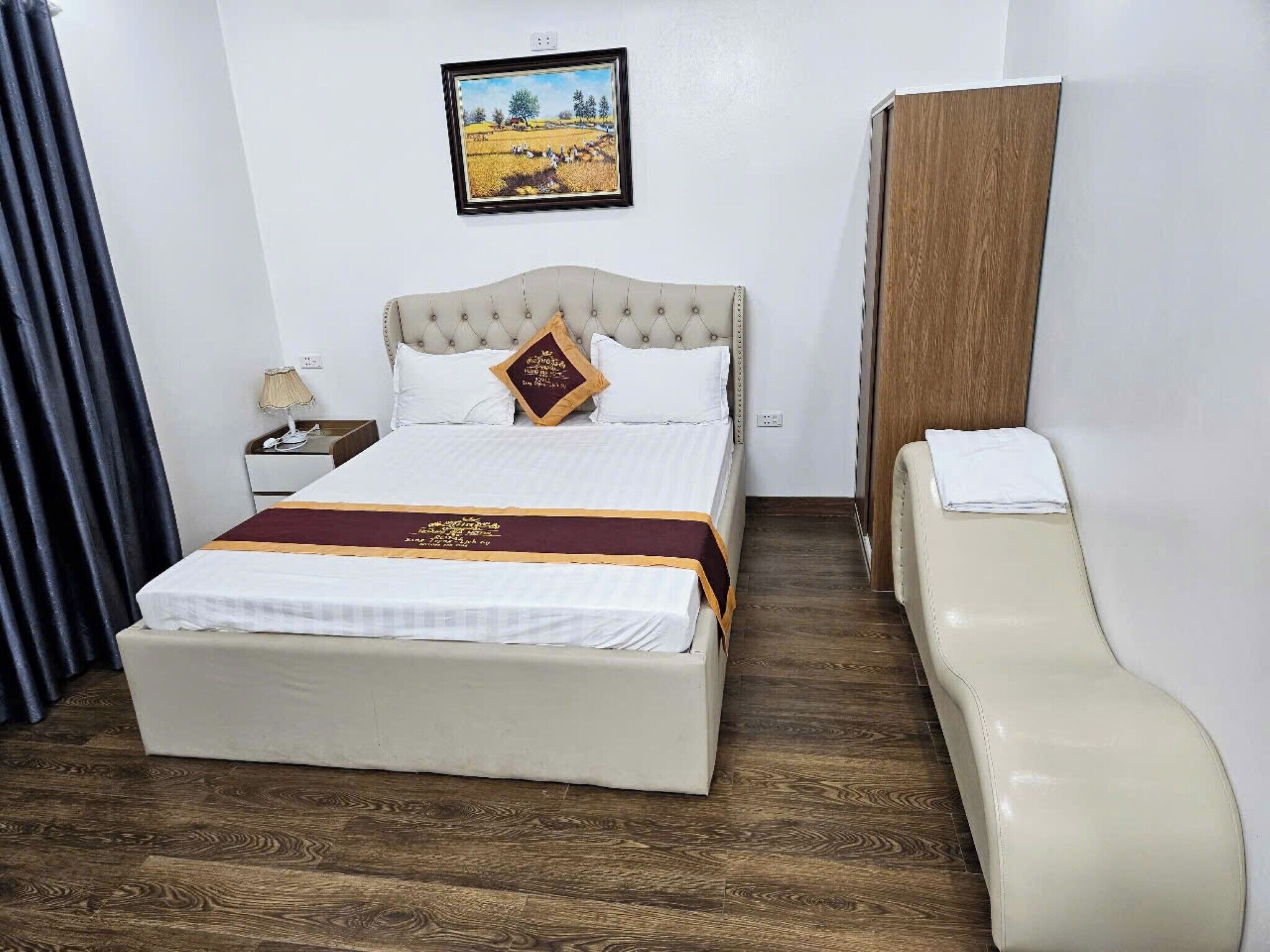 A hotel room with a large bed, a bedside table with a lamp, a wooden headboard, and a painting on the wall.