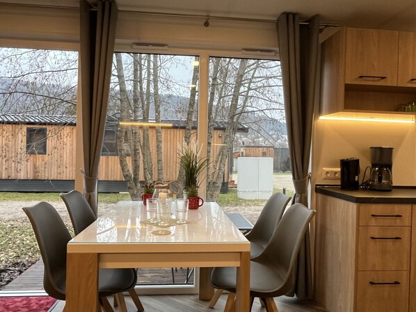 Enjoy meals at the large table surrounded by comfortable chairs, with a kitchen area nearby and a serene forest view through the large windows.