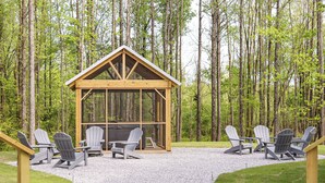 A wooden cabin with a glass front, surrounded by Adirondack chairs and a gravel path.