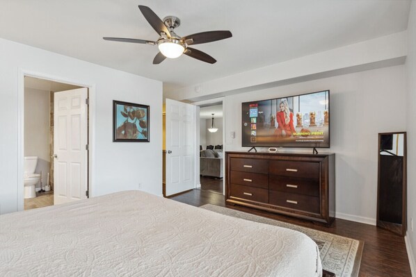 Une chambre avec un grand lit, un ventilateur de plafond, un téléviseur posé sur une commode en bois et une porte donnant sur une autre pièce.