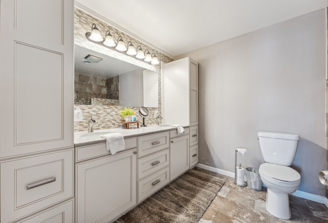 Une salle de bain avec un meuble-lavabo blanc, un grand miroir avec éclairage intégré, des toilettes et un tapis à motifs.