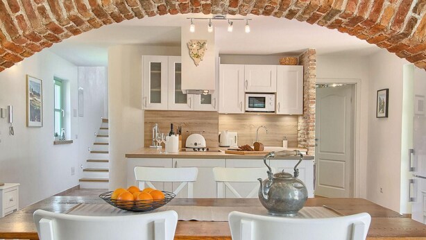 Une cuisine moderne avec des meubles blancs, un plan de travail en bois et une arche en briques. On y trouve une table à manger avec des chaises et un saladier rempli de fruits.