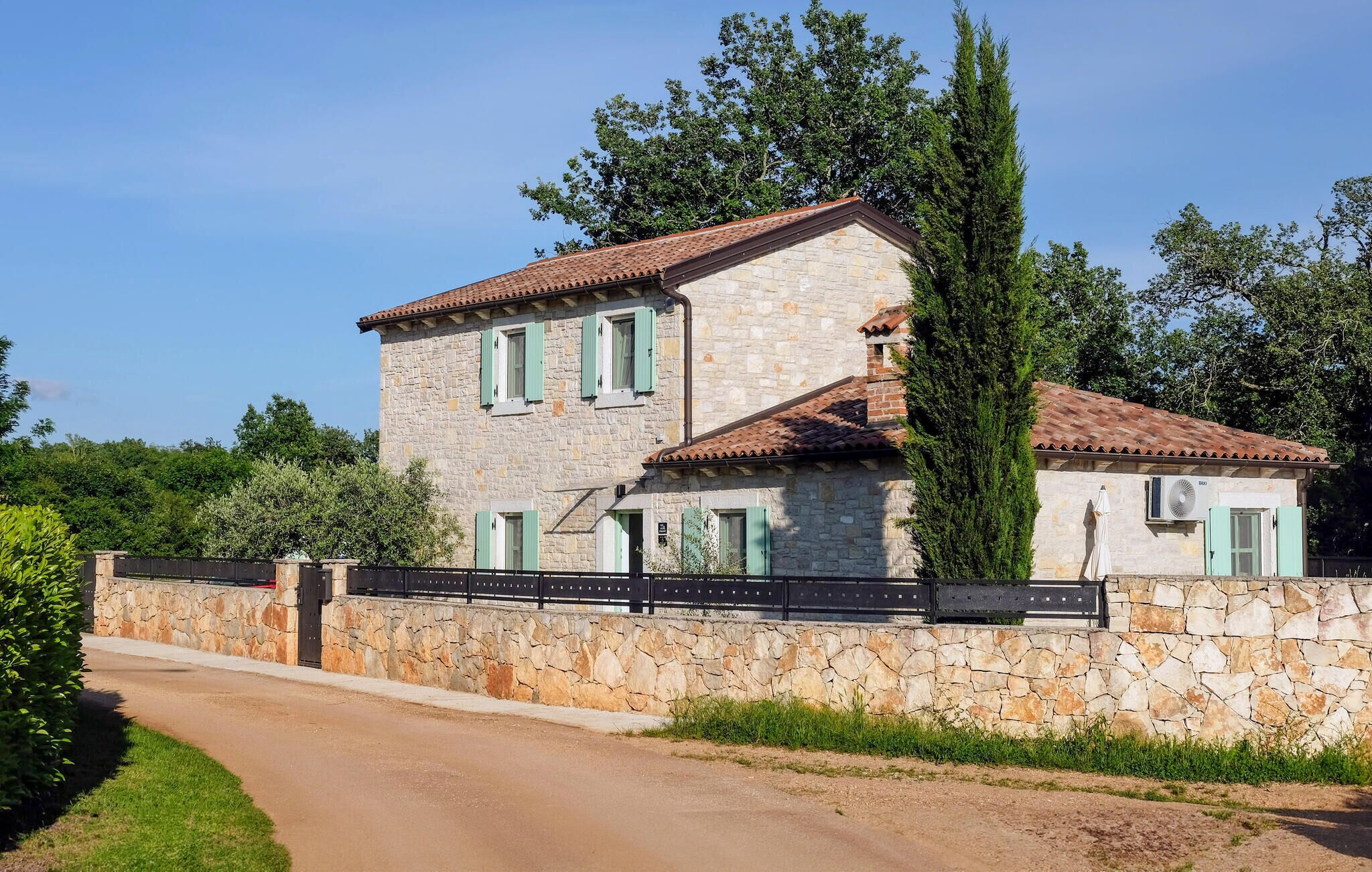 A two-story stone house with a tiled roof, green shutters, and a stone wall with a gate.