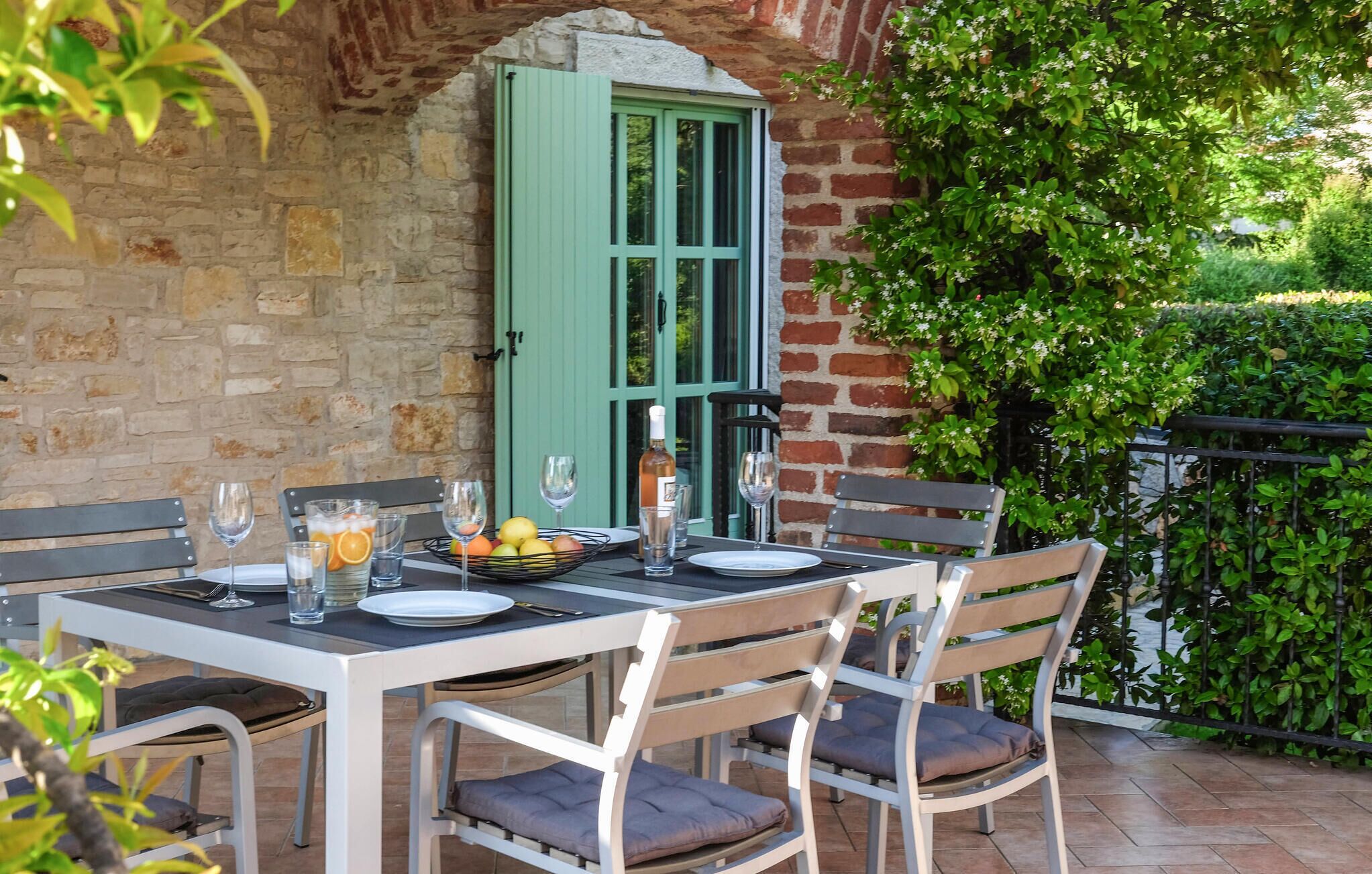 An outdoor dining area with a brick wall, green shutters, and a table set with plates, glasses, and a fruit bowl.
