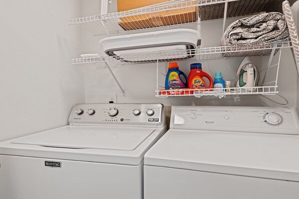 Ein Waschraum mit einer Maytag Waschmaschine und einem Trockner, einem Regal mit Waschmittelflaschen und einem Kleiderständer mit einem gemusterten Kleidungsstück.