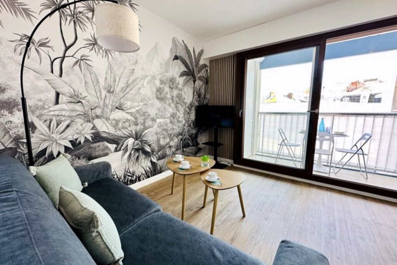 Ett modernt vardagsrum med en grå soffa, ett litet träbord med koppar, ett stort fönster med utsikt över omgivningen och en väggmålning med tropiska blad.