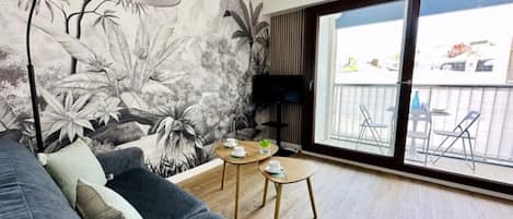 Ett modernt vardagsrum med en grå soffa, ett litet träbord med koppar, ett stort fönster med utsikt över omgivningen och en väggmålning med tropiska blad.