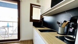 Eine moderne Küche mit schwarzem Arbeitsplatz, einer Kaffeemaschine und einem Set an Besteck. Ein Fenster bietet Blick auf einen Balkon und einen Tisch im Freien.