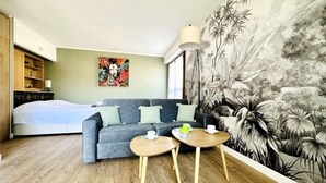 Ein modernes Zimmer mit einem grauen Sofa, zwei kleinen Holztischen, einem Bett mit weißer Bettdecke, einem Bücherregal und einem großen Blumenwandbild.