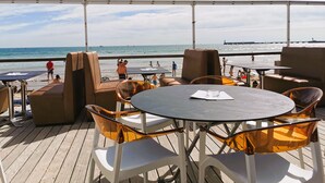 Ein Sitzbereich im Freien mit runden Tischen und Stühlen, mit Blick auf einen Strand, auf dem sich Menschen befinden, und einen Leuchtturm in der Ferne.