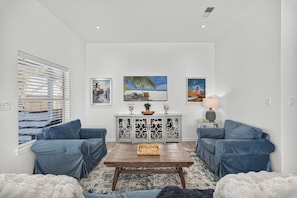 A living room with two blue sofas, a wooden coffee table, a cabinet with decorative items, and three framed paintings on the wall. - Private Heated Pool - Hot Tub - Golf Cart - Block to Beach - Fully Remodeled! (Destin)