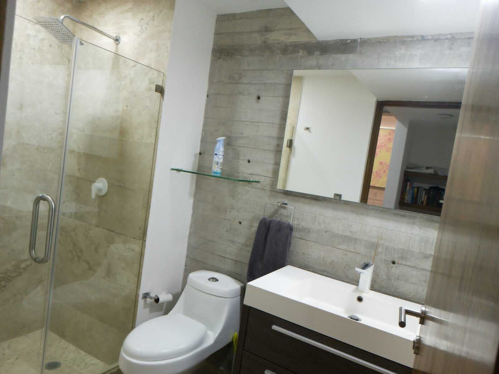 A bathroom with a glass shower enclosure, a white toilet, a vanity with a mirror, and a shelf with toiletries.