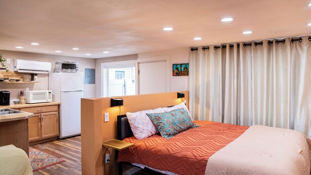 Een compacte hotelkamer met kitchenette, een bed met een decoratieve spreidek en uitzicht buiten door een raam met gordijnen.