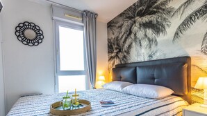 En pænt redt seng med et stribet tæppe, et sengebord med en lampe, og en væg med et palmetræsmaleri.