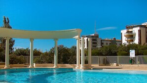 En swimmingpool med en hvid pergola, omgivet af hegn og grønt.