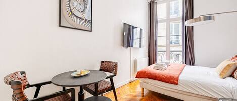 A hotel room with a bed, a round table with chairs, a TV, and a window with curtains.