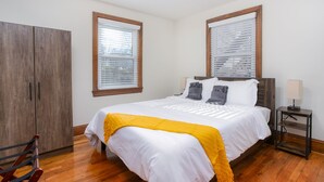 Un dormitorio con un armario de madera, una cama con ropa de cama blanca y un plaid amarillo, dos mesitas de noche con lámparas y una ventana con persianas.