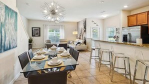 A modern kitchen with a refrigerator, a dining area with a glass table and chairs, and a living area with a sofa and a coffee table. - 5BD Paradise Palms Resort Near Parks (PP 8976) (Kissimmee)