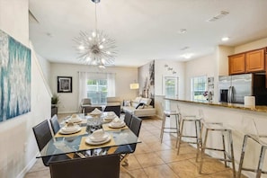 Una cocina moderna con refrigerador, un comedor con mesa y sillas de vidrio, y una sala de estar con sofá y mesa de centro.