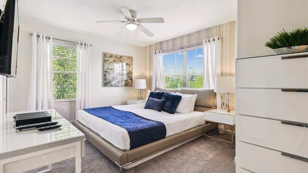 A hotel room with a bed, a desk with a computer, a ceiling fan, and a window with curtains. - 5BD Paradise Palms Resort Near Parks (PP 8976) (Kissimmee)