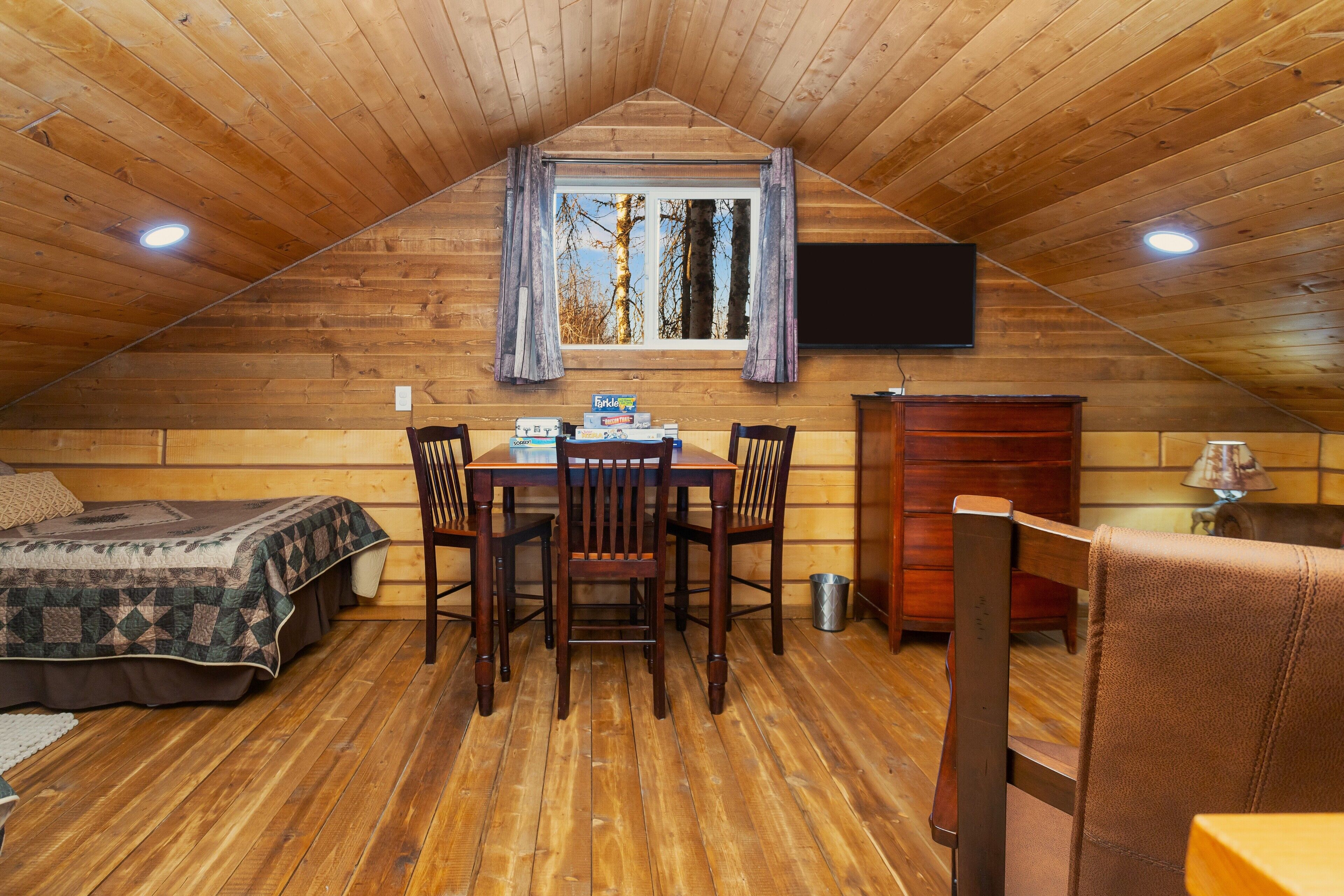 Un chalet confortable avec un intérieur en bois, une table à manger avec des chaises, un lit avec une couverture et un téléviseur fixé au mur.