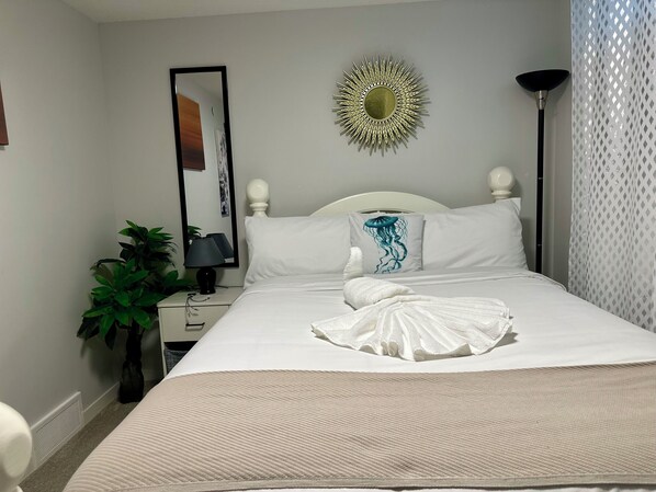 A neatly made bed with a white comforter and a decorative pillow, a bedside lamp, a mirror, and a potted plant. - Global Family Canada (Calgary)