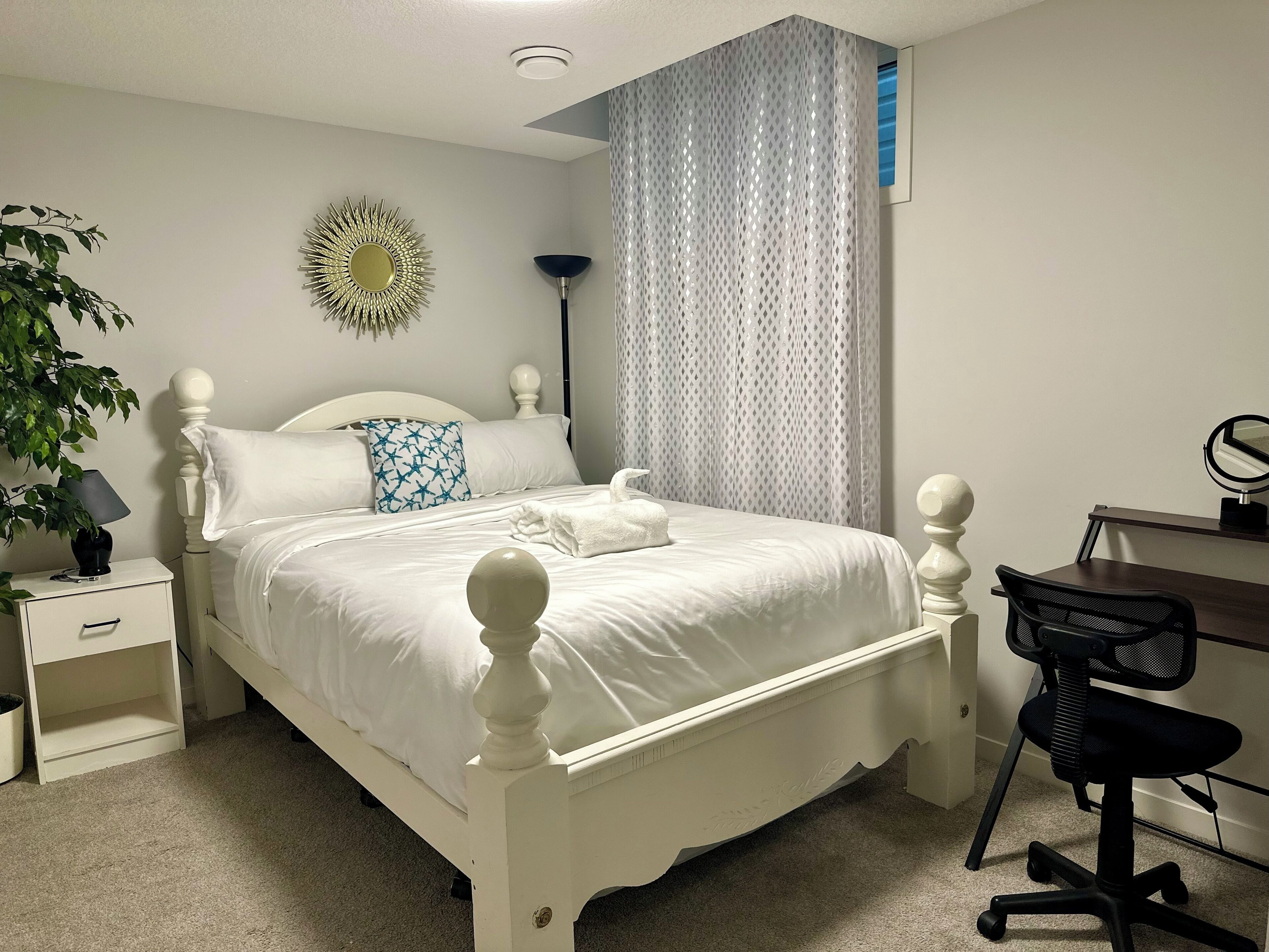 A neatly made bed with white bedding, a bedside lamp, a decorative mirror, and a desk with a chair.