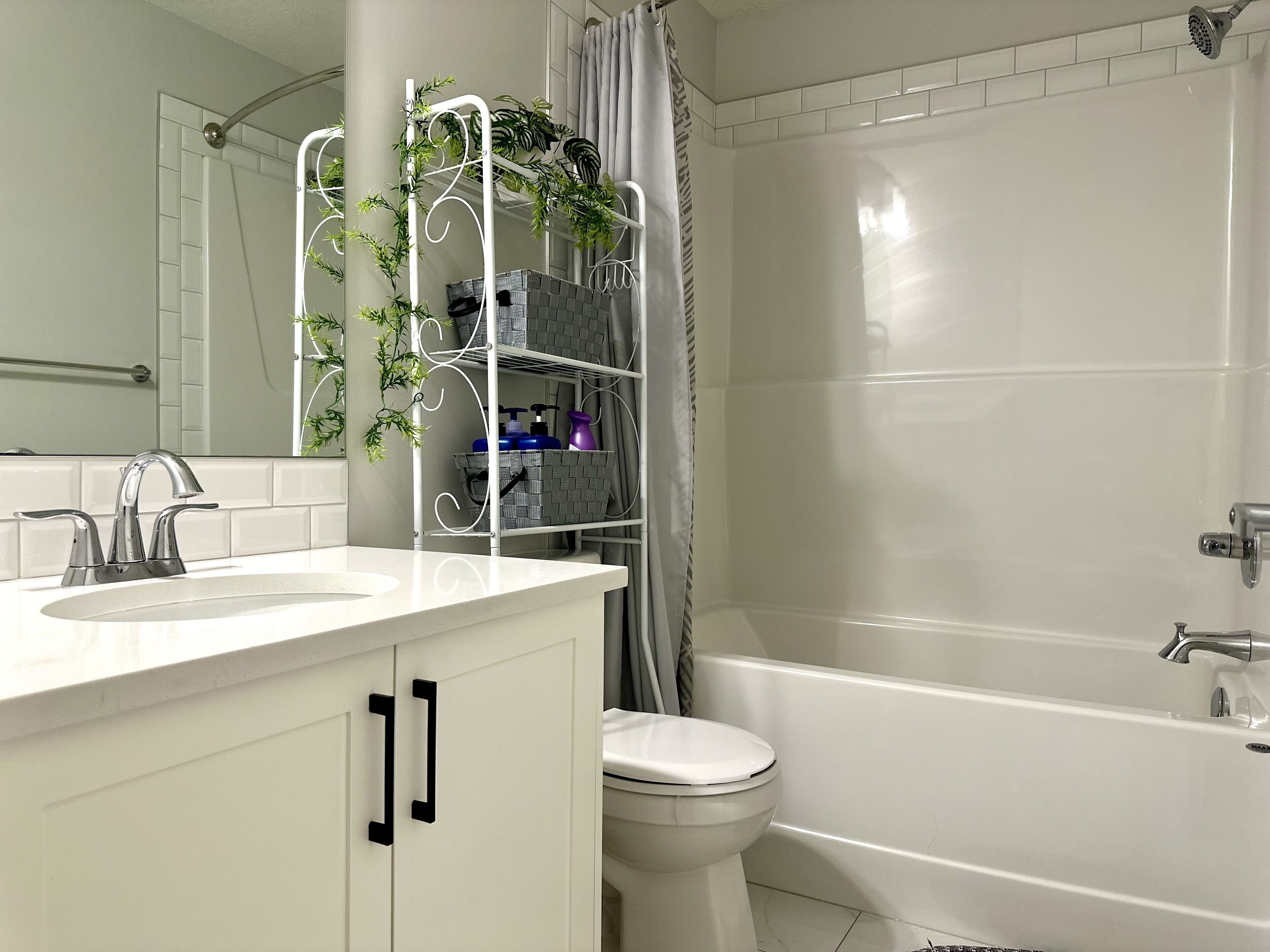 Un baño con lavamanos blanco, inodoro y bañera. Hay una repisa con plantas y artículos de aseo.
