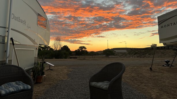 En campingvogn med mærket 'Outback by Sydney', et grusområde med to fletstole, og en himmel i solnedgang.