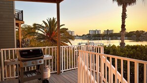 Una terraza de madera con una parrilla y un tanque de gas, con vista a un cuerpo de agua donde se ven barcos y edificios al fondo.