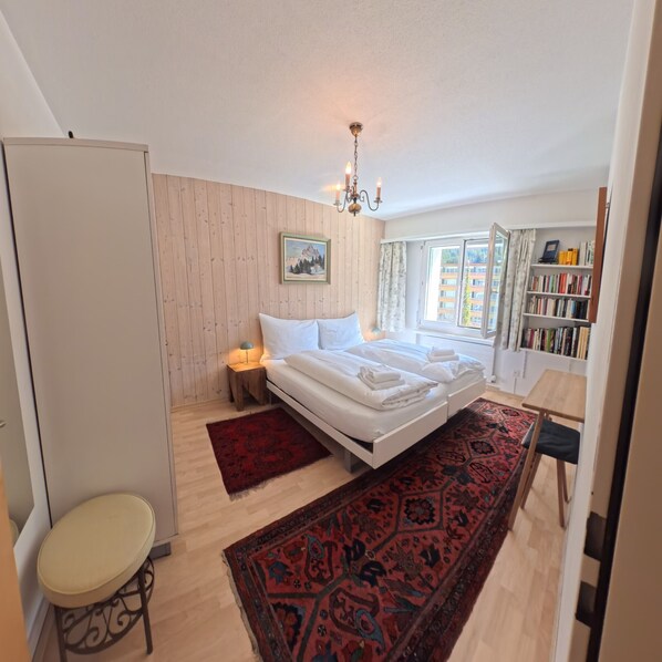 A bedroom with a bed, a nightstand, a chair, a bookshelf, and a window with a view.