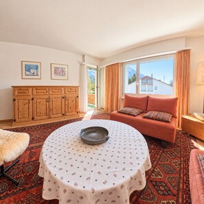 A cozy living room with a round table, a sofa with cushions, a wooden cabinet, and a view of the outdoors through a large window. - Central base for biking and skiing (Lenzerheide/Lai)