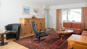 Una sala de estar con un aparador de madera, una silla negra, una mesa redonda con fruta, un sofá rojo y una ventana con cortinas.