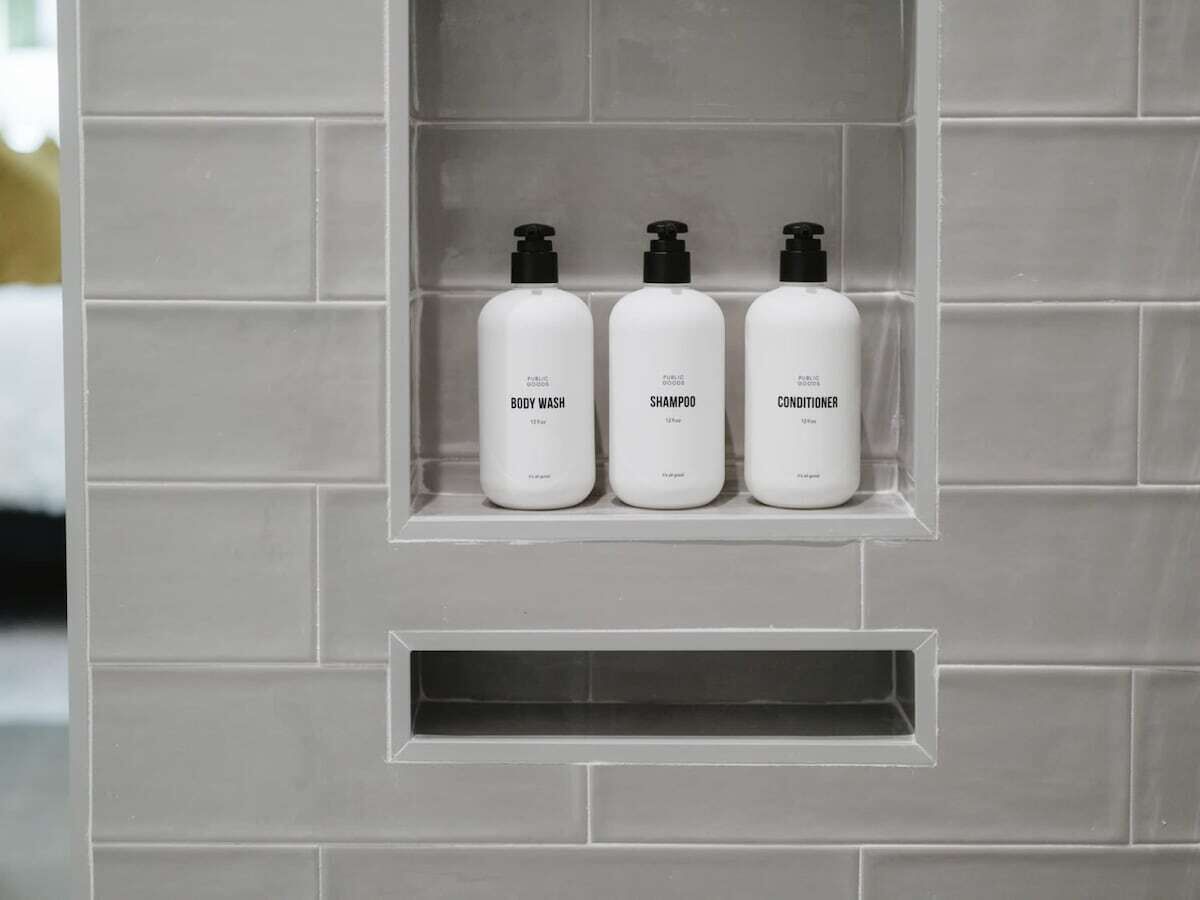 Tres botellas blancas de jabón corporal, champú y acondicionador sobre una pared de baldosas.