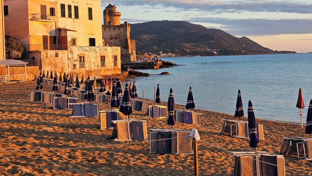 Une plage bordée de rangées de chaises longues et de parasols, un bâtiment historique doté d’une tour, et une vue sur la mer et les collines en arrière-plan.