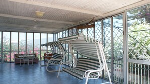 A covered patio area with striped lounge chairs and a sofa, large windows offering views of greenery and a building.
