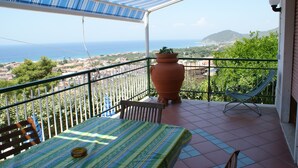 A balcony with a table, a chair, a potted plant, and a view of the sea and hills.