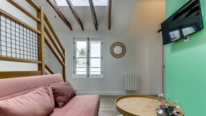 Un salon avec un canapé rose, une table basse ronde en bois, un téléviseur fixé sur un mur vert et une fenêtre donnant sur des immeubles.