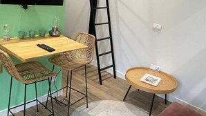 Une petite pièce meublée, avec une table en bois, deux chaises en osier, une petite table ronde, un canapé marron, une échelle et un téléviseur à écran plat fixé au mur.