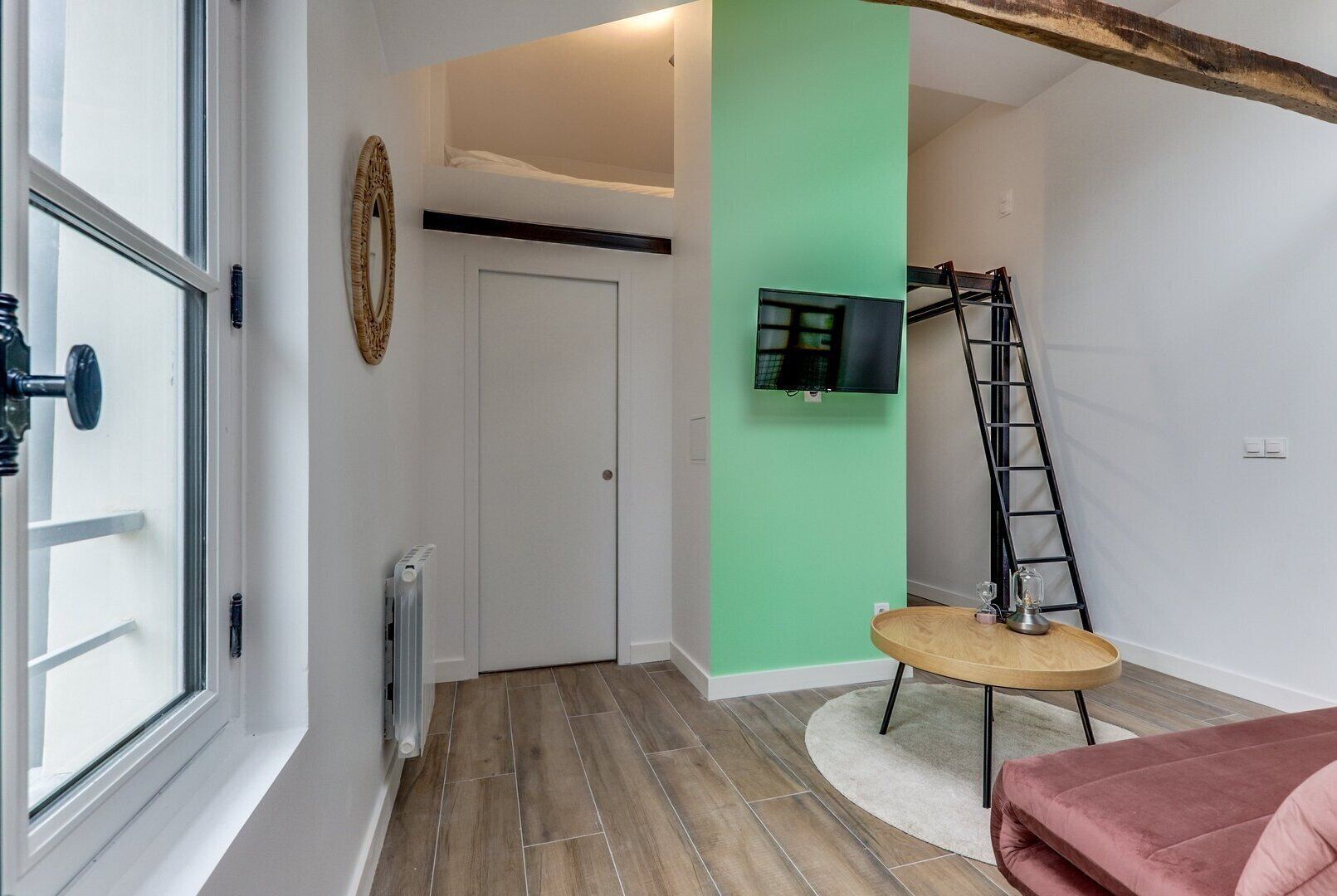 Una habitación con suelo de madera, una mesa redonda con superficie de vidrio, un sofá rosa, una escalera y un televisor fijado a la pared.
